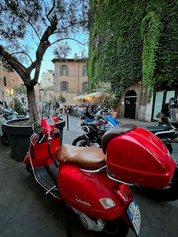 A vibrant red scooter is parked on a cobblestone street near a building covered in lush green ivy. The street is adorned with several other parked motorcycles, and there is a tree in the foreground. People can be seen sitting at outdoor tables under a canopy with lights, indicating a café or restaurant setting in the background. The buildings are traditional and have an old-world charm.