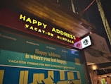 The image features a storefront at night, prominently displaying a sign with the words 'Happy Address' and 'Vacation Rentals' in bright yellow letters on a red background. Below, a large advertisement with the slogan 'Happy Address is where you feel happy' is visible, alongside logos of companies such as TripAdvisor, Airbnb, and Expedia.