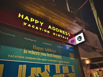 The image features a storefront at night, prominently displaying a sign with the words 'Happy Address' and 'Vacation Rentals' in bright yellow letters on a red background. Below, a large advertisement with the slogan 'Happy Address is where you feel happy' is visible, alongside logos of companies such as TripAdvisor, Airbnb, and Expedia.