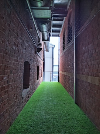 A narrow corridor with exposed red brick walls on each side, featuring a bright green artificial turf floor. Overhead, industrial metal pipes and ductwork are visible against the ceiling, creating a contrast with the natural texture of the walls. The corridor ends with a metal railing, allowing a view of a larger structure outside.