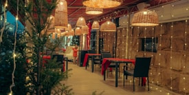 A cozy outdoor dining area adorned with warm fairy lights and wicker lampshades, creating an inviting ambiance. Tables covered with red cloths and black chairs are neatly arranged. Evergreen plants and a person dressed in a warm coat add to the wintery, festive atmosphere.