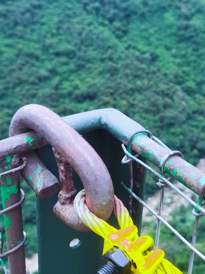 A metal U-bolt clamp secured to a section of wire mesh fence with visible signs of rust and peeling green paint. The background shows a blurred green landscape, possibly trees or shrubs. A bright yellow component of the clamp contrasts with the otherwise muted and earthy tones.