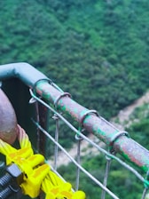 A close-up view of a sturdy metal railing attached to wire cables, with a background of lush greenery and a river below. The railing shows signs of weathering, with patches of green paint on rusted metal. Bright yellow cable clamps are visibly attached to the cables, and the scene has a serene and natural setting.