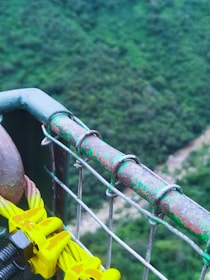 A close-up view of a sturdy metal railing attached to wire cables, with a background of lush greenery and a river below. The railing shows signs of weathering, with patches of green paint on rusted metal. Bright yellow cable clamps are visibly attached to the cables, and the scene has a serene and natural setting.