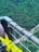 A close-up view of a sturdy metal railing attached to wire cables, with a background of lush greenery and a river below. The railing shows signs of weathering, with patches of green paint on rusted metal. Bright yellow cable clamps are visibly attached to the cables, and the scene has a serene and natural setting.