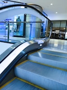 An indoor escalator ascending in a modern shopping mall environment with a glass safety barrier on one side. In the background, there is a display window featuring a mannequin dressed in casual clothing. The scene is well-lit, with bright overhead lights and a sleek, contemporary design.