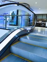 An indoor escalator ascending in a modern shopping mall environment with a glass safety barrier on one side. In the background, there is a display window featuring a mannequin dressed in casual clothing. The scene is well-lit, with bright overhead lights and a sleek, contemporary design.