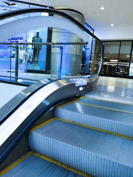 An indoor escalator ascending in a modern shopping mall environment with a glass safety barrier on one side. In the background, there is a display window featuring a mannequin dressed in casual clothing. The scene is well-lit, with bright overhead lights and a sleek, contemporary design.