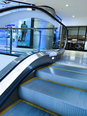 An indoor escalator ascending in a modern shopping mall environment with a glass safety barrier on one side. In the background, there is a display window featuring a mannequin dressed in casual clothing. The scene is well-lit, with bright overhead lights and a sleek, contemporary design.