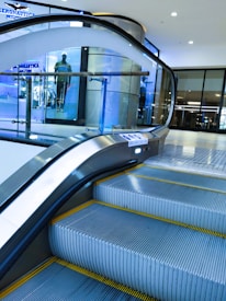 An indoor escalator ascending in a modern shopping mall environment with a glass safety barrier on one side. In the background, there is a display window featuring a mannequin dressed in casual clothing. The scene is well-lit, with bright overhead lights and a sleek, contemporary design.