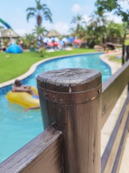 A close-up view of a wooden railing with visible grain and texture. In the background, a swimming pool with a person on a yellow inflatable float is seen, surrounded by lush green grass and tall palm trees. The scene is set in a bright, sunny outdoor location with a relaxed atmosphere.