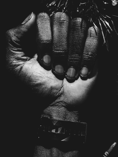 A close-up black and white image of a clenched human fist. The fist is prominently centered in the frame, with detailed textures and lines visible on the skin. The lighting creates dramatic shadows and highlights, emphasizing the contours of the hand. A bracelet is visible on the wrist, adding a touch of detail to the composition.