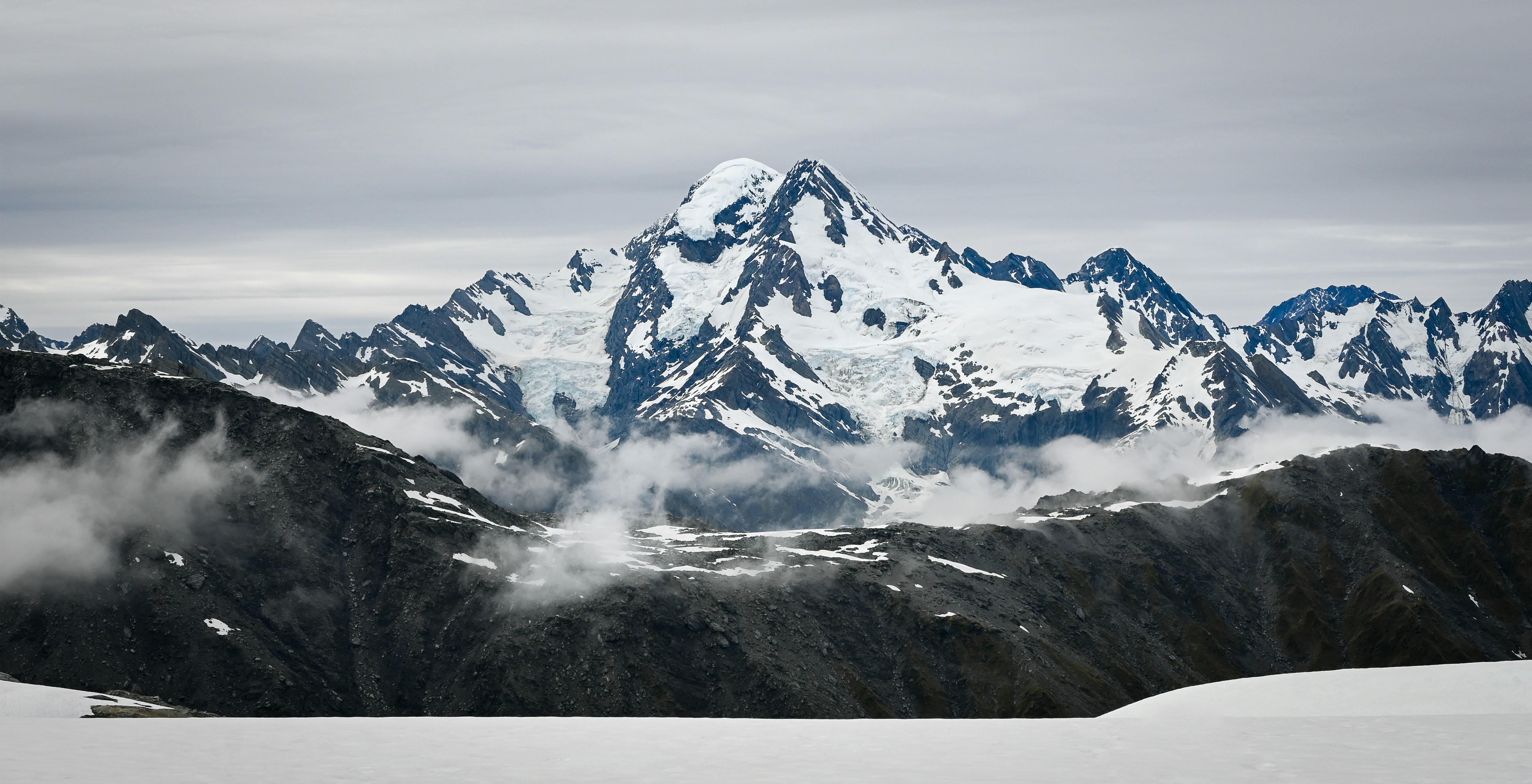 a snow covered mountain range with clouds in the foreground, In a seemingly endless abyss of mountains and glaciers, a small flat snowfield with near-perfect picturesque views lies where this photo was taken.