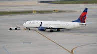 A commercial airplane is being towed by a small tow vehicle on an airport tarmac. The aircraft, with Delta Airlines branding, is positioned on a large paved surface with visible taxiway markings. The airplane and tow vehicle create a sense of movement and readiness.