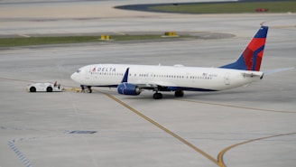 A commercial airplane is being towed by a small tow vehicle on an airport tarmac. The aircraft, with Delta Airlines branding, is positioned on a large paved surface with visible taxiway markings. The airplane and tow vehicle create a sense of movement and readiness.