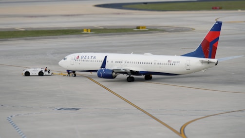 A commercial airplane is being towed by a small tow vehicle on an airport tarmac. The aircraft, with Delta Airlines branding, is positioned on a large paved surface with visible taxiway markings. The airplane and tow vehicle create a sense of movement and readiness.