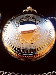 An ornate gold pocket watch with an intricate engraved design on the cover, featuring a shield-like emblem at the center. The watch is positioned against a dark background, highlighting its reflective surface and detailed craftsmanship.