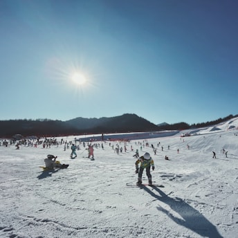 A snowy landscape with numerous people engaged in skiing and snowboarding activities. The bright sun casts long shadows on the snow, and a backdrop of distant mountains is visible under a clear blue sky.