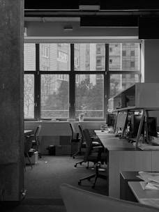 A monochrome image of an empty office space with several chairs and desks arranged neatly. Computer monitors are placed on the desks, with various office supplies scattered around. Large windows at the back provide a view of the outside buildings and trees. The atmosphere appears quiet and serene, with natural light coming through the windows, illuminating the workspace.
