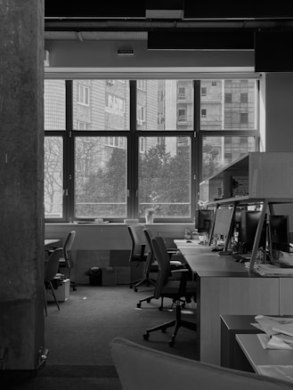 A monochrome image of an empty office space with several chairs and desks arranged neatly. Computer monitors are placed on the desks, with various office supplies scattered around. Large windows at the back provide a view of the outside buildings and trees. The atmosphere appears quiet and serene, with natural light coming through the windows, illuminating the workspace.