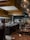 A modern and stylish restaurant interior with an open kitchen and chefs actively preparing dishes. The dining area features wooden tables set with glasses and candles, providing a warm and inviting atmosphere. Large pendant lights hang from the ceiling, and there is a wall with stacked wood logs, adding a rustic touch.