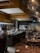 A modern and stylish restaurant interior with an open kitchen and chefs actively preparing dishes. The dining area features wooden tables set with glasses and candles, providing a warm and inviting atmosphere. Large pendant lights hang from the ceiling, and there is a wall with stacked wood logs, adding a rustic touch.