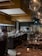 A modern and stylish restaurant interior with an open kitchen and chefs actively preparing dishes. The dining area features wooden tables set with glasses and candles, providing a warm and inviting atmosphere. Large pendant lights hang from the ceiling, and there is a wall with stacked wood logs, adding a rustic touch.