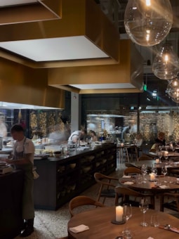 A modern and stylish restaurant interior with an open kitchen and chefs actively preparing dishes. The dining area features wooden tables set with glasses and candles, providing a warm and inviting atmosphere. Large pendant lights hang from the ceiling, and there is a wall with stacked wood logs, adding a rustic touch.