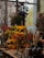A vibrant display of various flowers and plants, including red, yellow, and green floral arrangements. Large dried plants and berries are visible in the background, creating a rustic ambiance. Glass vases and decorative gourds are arranged on a wooden table, accompanied by other garden and home accessories.