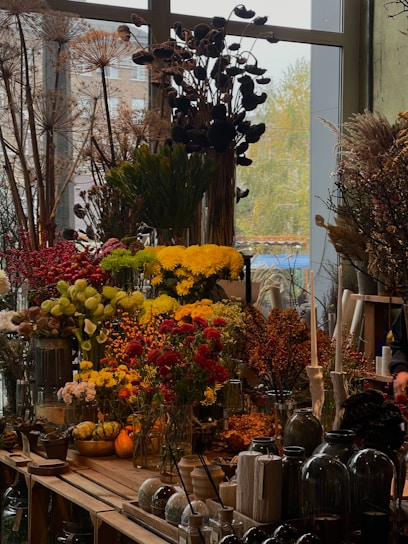 A vibrant display of various flowers and plants, including red, yellow, and green floral arrangements. Large dried plants and berries are visible in the background, creating a rustic ambiance. Glass vases and decorative gourds are arranged on a wooden table, accompanied by other garden and home accessories.