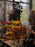 A vibrant display of various flowers and plants, including red, yellow, and green floral arrangements. Large dried plants and berries are visible in the background, creating a rustic ambiance. Glass vases and decorative gourds are arranged on a wooden table, accompanied by other garden and home accessories.