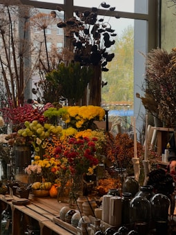 A vibrant display of various flowers and plants, including red, yellow, and green floral arrangements. Large dried plants and berries are visible in the background, creating a rustic ambiance. Glass vases and decorative gourds are arranged on a wooden table, accompanied by other garden and home accessories.
