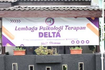 A large banner with the text 'Lembaga Psikologi Terapan DELTA' is displayed prominently. A logo and contact information are included, featuring a website, email address, and phone number. Below the banner, there are two potted plants placed on a black structure.