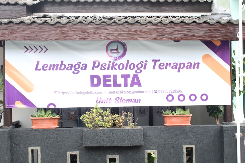 A large banner with the text 'Lembaga Psikologi Terapan DELTA' is displayed prominently. A logo and contact information are included, featuring a website, email address, and phone number. Below the banner, there are two potted plants placed on a black structure.