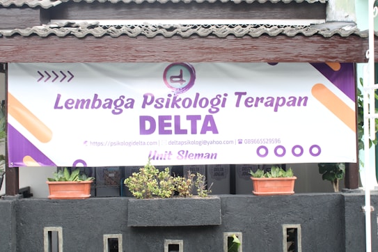 A large banner with the text 'Lembaga Psikologi Terapan DELTA' is displayed prominently. A logo and contact information are included, featuring a website, email address, and phone number. Below the banner, there are two potted plants placed on a black structure.
