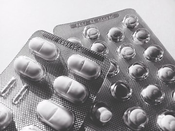 Blister packs containing tablets and pills, arranged overlapping on a flat surface. The image is in black and white, highlighting texture and form.