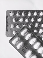 Two blister packs containing pills are placed on a light surface. The packs are arranged at slight angles to each other, with one overlapping the top left corner of the other. The lighting creates shadows that emphasize the texture of the blister packs and the shapes of the individual pill compartments.