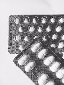 Two blister packs containing pills are placed on a light surface. The packs are arranged at slight angles to each other, with one overlapping the top left corner of the other. The lighting creates shadows that emphasize the texture of the blister packs and the shapes of the individual pill compartments.