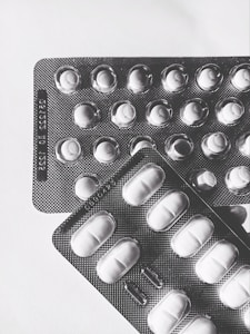 Two blister packs containing pills are placed on a light surface. The packs are arranged at slight angles to each other, with one overlapping the top left corner of the other. The lighting creates shadows that emphasize the texture of the blister packs and the shapes of the individual pill compartments.