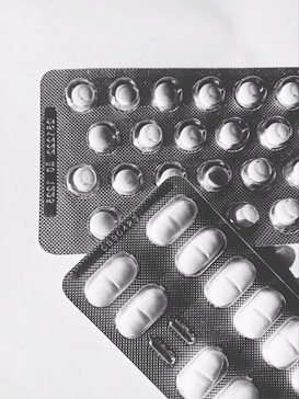 Two blister packs containing pills are placed on a light surface. The packs are arranged at slight angles to each other, with one overlapping the top left corner of the other. The lighting creates shadows that emphasize the texture of the blister packs and the shapes of the individual pill compartments.