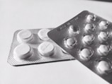 Two blister packs containing round, white tablets are placed on a flat surface. The tablets have a cross imprint on them, and the packs overlap each other slightly.