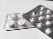 Two blister packs containing round, white tablets are placed on a flat surface. The tablets have a cross imprint on them, and the packs overlap each other slightly.