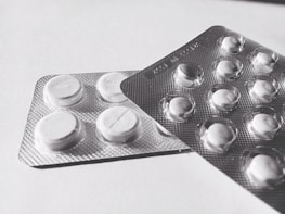 Two blister packs containing round, white tablets are placed on a flat surface. The tablets have a cross imprint on them, and the packs overlap each other slightly.