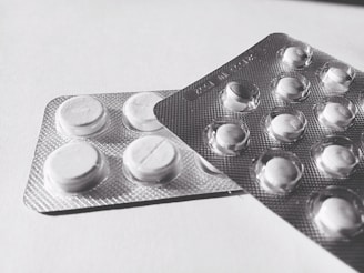 Two blister packs containing round, white tablets are placed on a flat surface. The tablets have a cross imprint on them, and the packs overlap each other slightly.