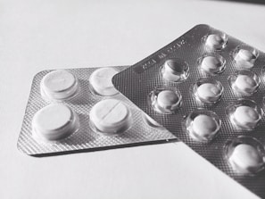 Two blister packs containing round, white tablets are placed on a flat surface. The tablets have a cross imprint on them, and the packs overlap each other slightly.