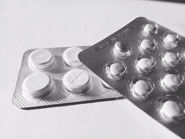 Two blister packs containing round, white tablets are placed on a flat surface. The tablets have a cross imprint on them, and the packs overlap each other slightly.