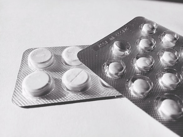 Two blister packs containing round, white tablets are placed on a flat surface. The tablets have a cross imprint on them, and the packs overlap each other slightly.