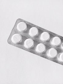 A silver blister pack containing white round pills, placed on a clean, plain background. The packaging is textured and reflective, and each pill is separated in its own compartment.