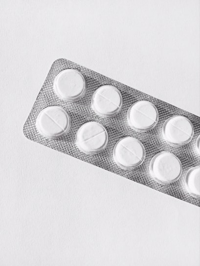 A silver blister pack containing white round pills, placed on a clean, plain background. The packaging is textured and reflective, and each pill is separated in its own compartment.