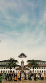 A large group of people is gathered in front of a historic building with unique architectural features, including a central tower. The Indonesian flag is raised above the building, and palm trees are situated on either side. The sky is blue with some clouds.
